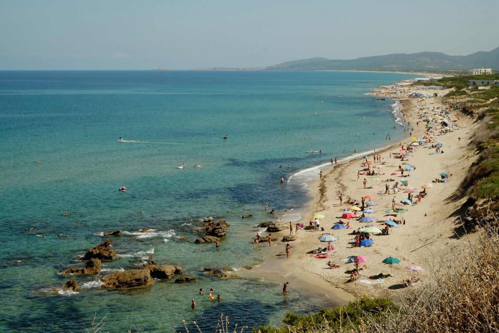 Langer Sandstrand auf Sardinien mit türkisfarbenem Meer, Sonnenbadenden, bunten Sonnenschirmen und klarem Sommerhimmel.