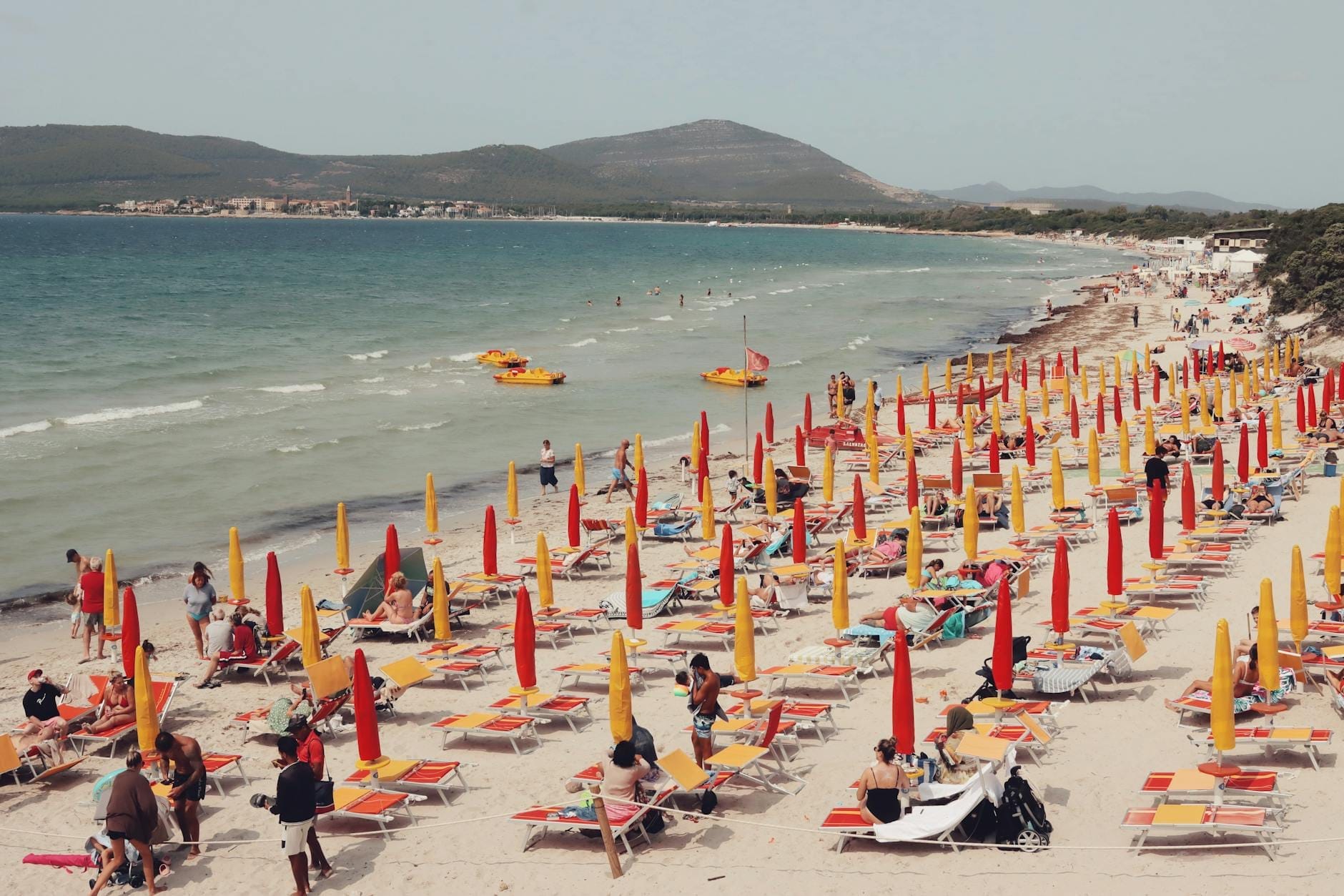 Belebter sardischer Strand mit bunten Sonnenschirmen und Liegestühlen, Menschen genießen das Meer mit malerischen Bergen im Hintergrund.