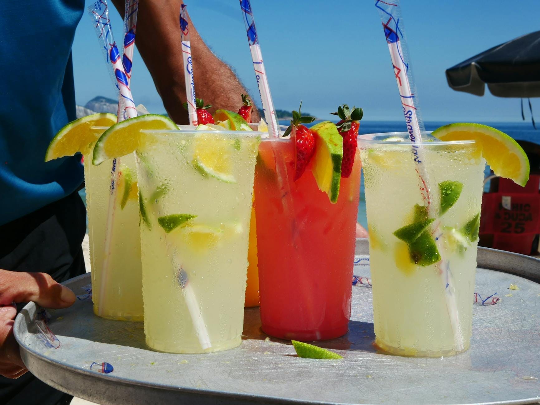 Nahaufnahme von Plastikbechern mit bunten alkoholfreien Getränken, garniert mit Zitrone und Früchten auf einem Tablett am Strand.