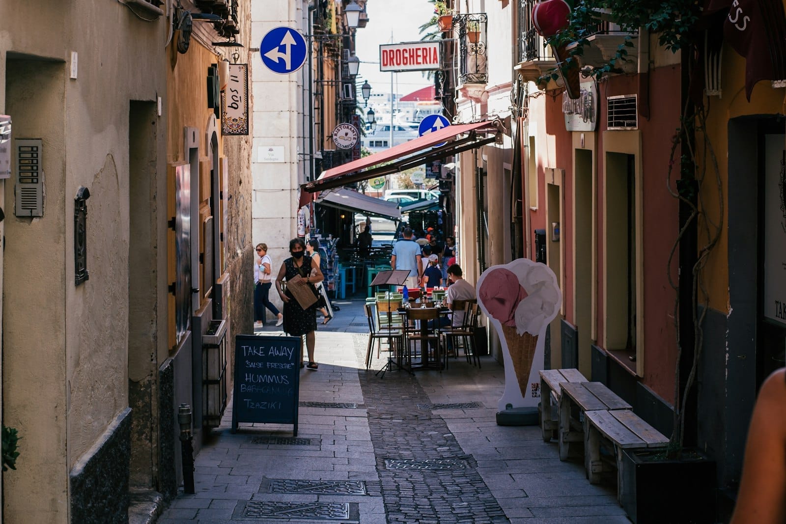 Enge, lebhafte sardische Straße mit kleinen Tischen, im Freien sitzenden Menschen, Restaurantschildern und typischen mediterranen Gebäuden.