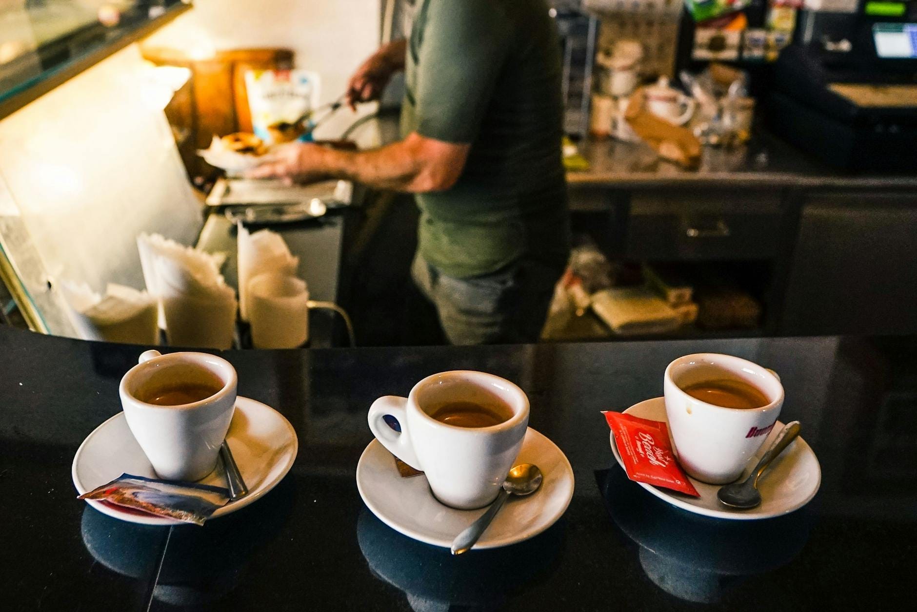 Drei Espressotassen auf dem Tresen eines Cafés mit einem Barista, der im Hintergrund Getränke zubereitet, und der italienischen Kaffeekultur.