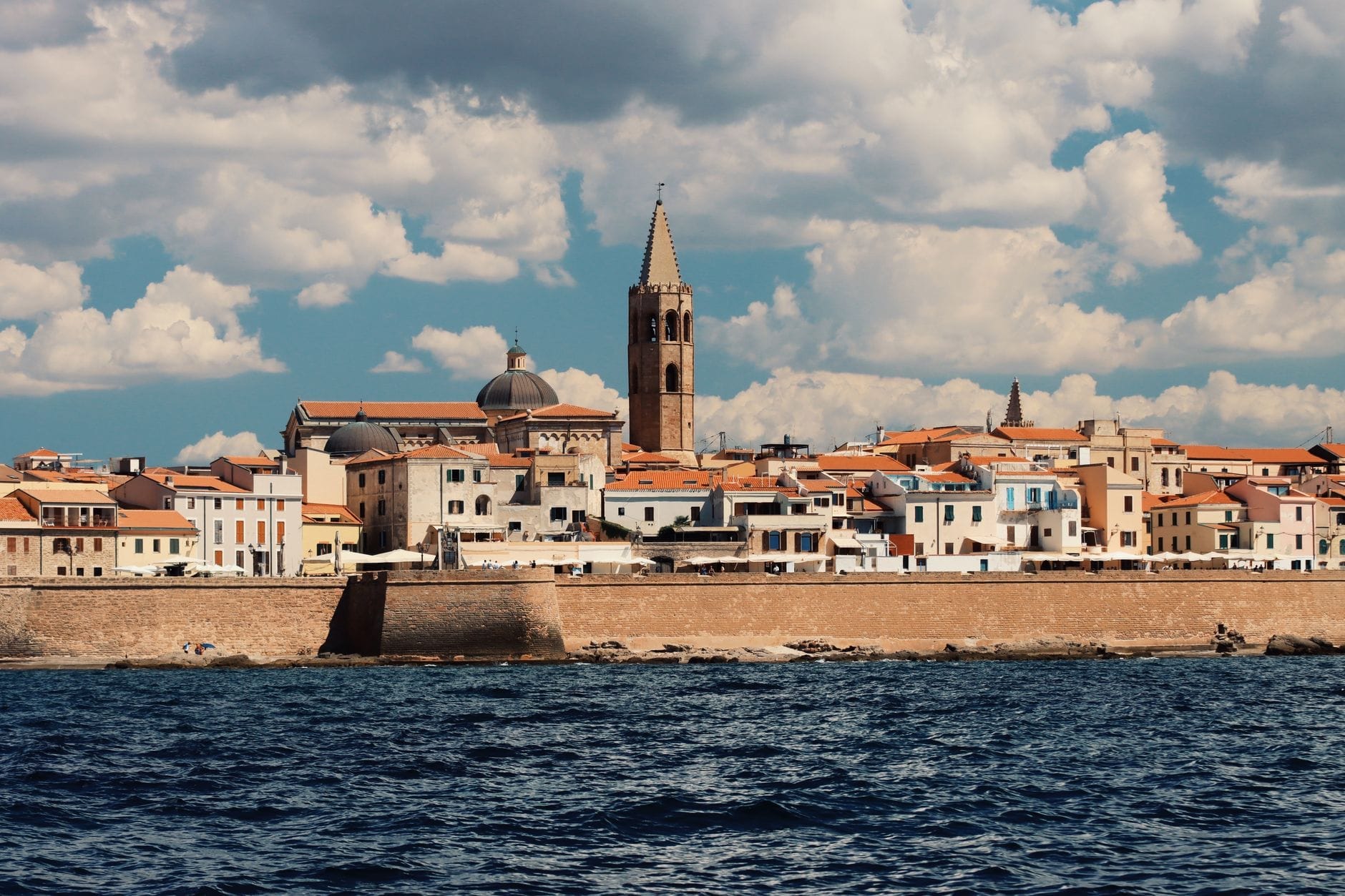 Blick auf eine historische sardische Küstenstadt mit bunten Gebäuden, einem markanten Kirchturm und Verteidigungsmauern am Meer unter leicht bewölktem Himmel.