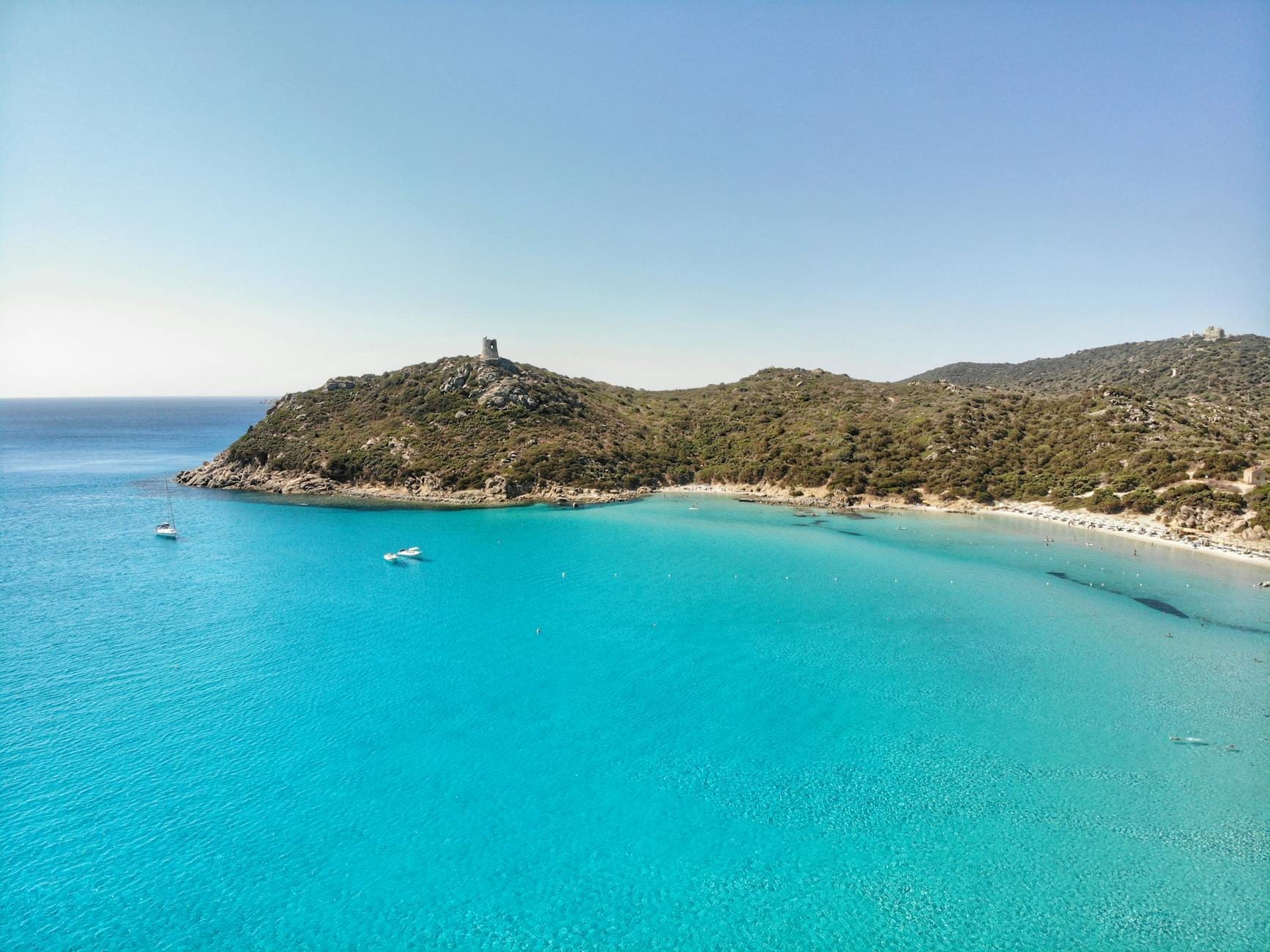 Luftaufnahme einer türkisfarbenen Bucht mit klarem Wasser, Booten und einem Küstenwachturm im Südosten Sardiniens in der Nähe von Villasimius.