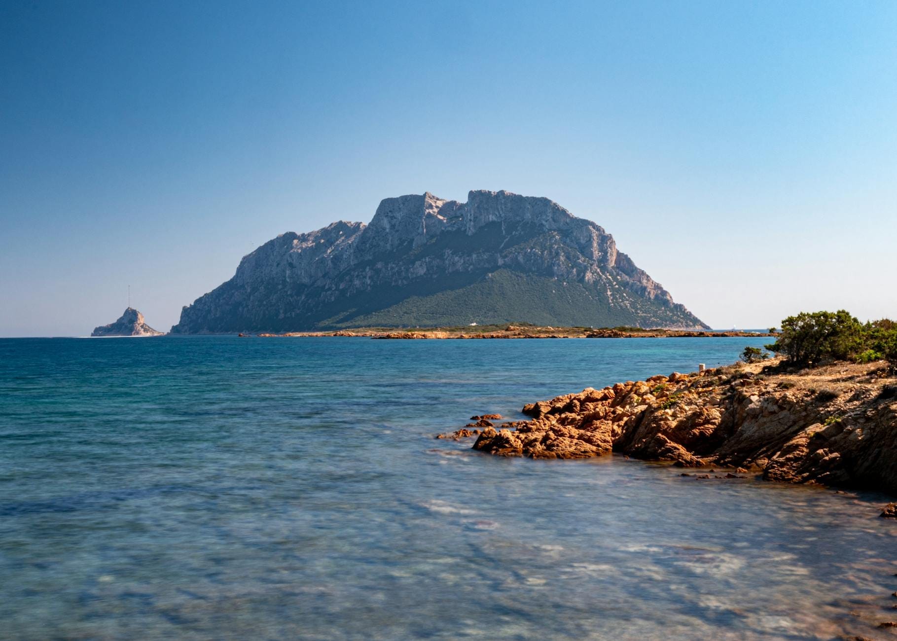Klarer Blick auf die Isola Tavolara, die aus dem türkisfarbenen Meer ragt, von einer felsigen Küste im Nordosten Sardiniens unter einem strahlend blauen Himmel.