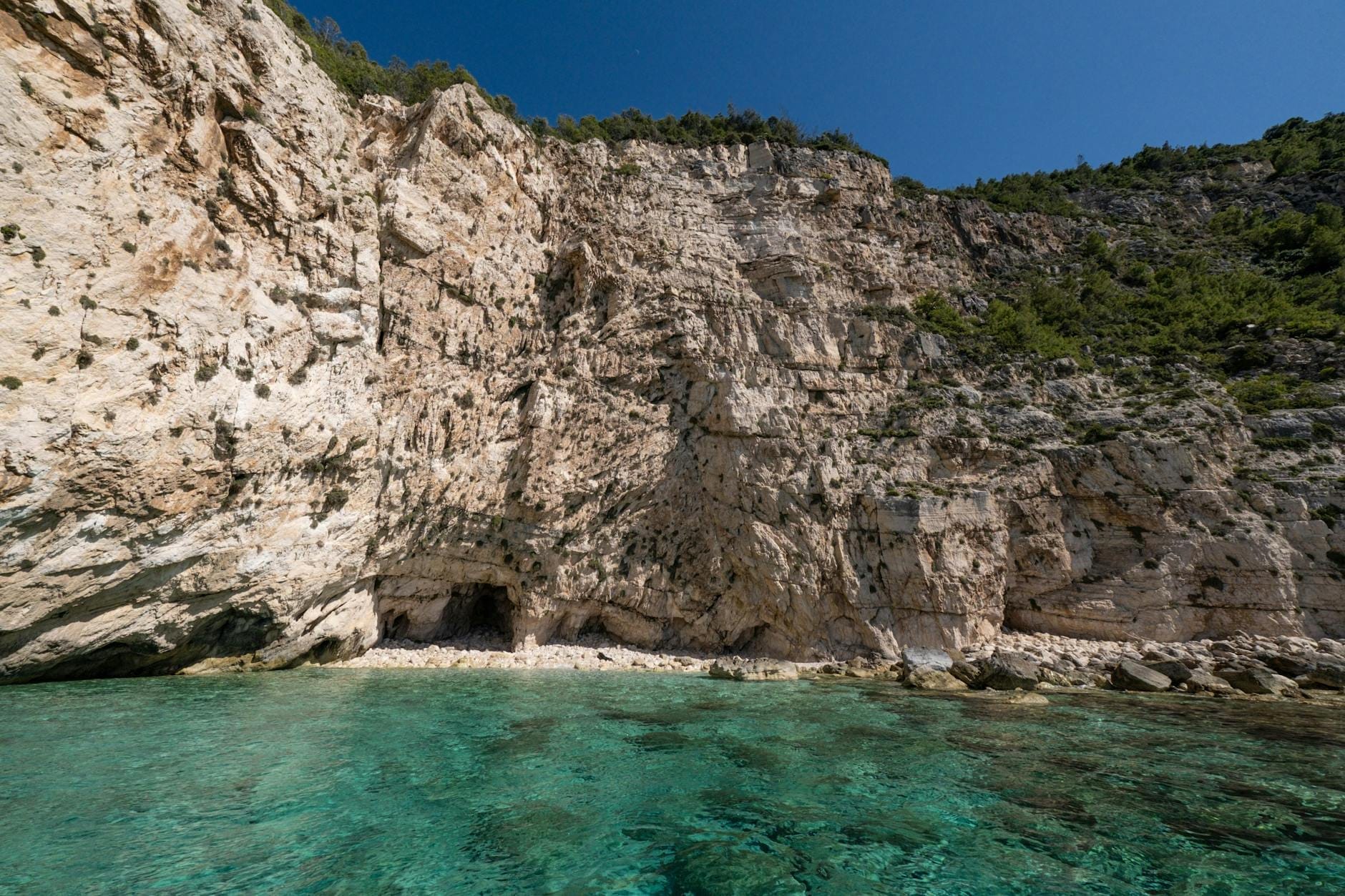Türkisfarbenes Wasser mit steilen Kalksteinklippen und kleinen Meereshöhlen entlang der Küste Sardiniens unter einem klaren blauen Himmel.