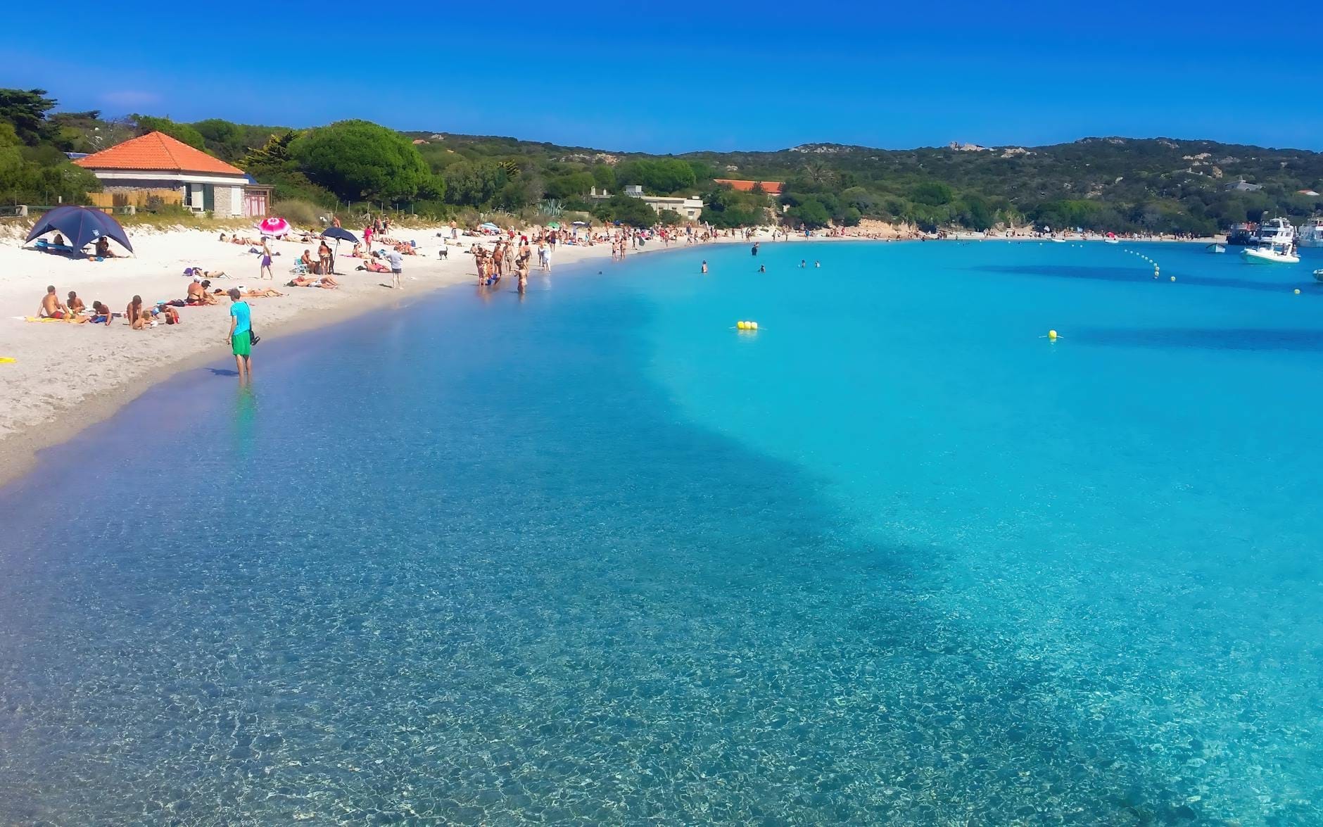 Wunderschöner Strand auf Sardinien mit klarem türkisfarbenem Wasser, weißem Sand, vereinzelten entspannenden Menschen und Booten in der Ferne an einem sonnigen Tag.