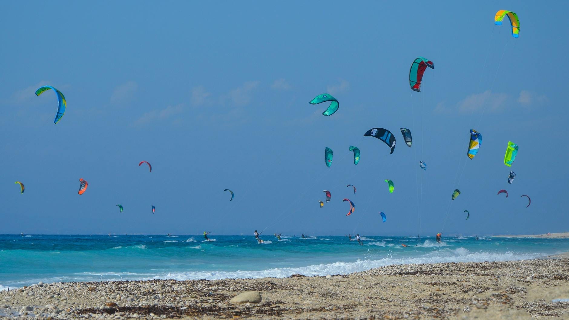 Dutzende bunte Kitesurf-Drachen füllen den Himmel über türkisfarbenem Wasser und einem Kiesstrand an einem sonnigen Tag auf Sardinien.