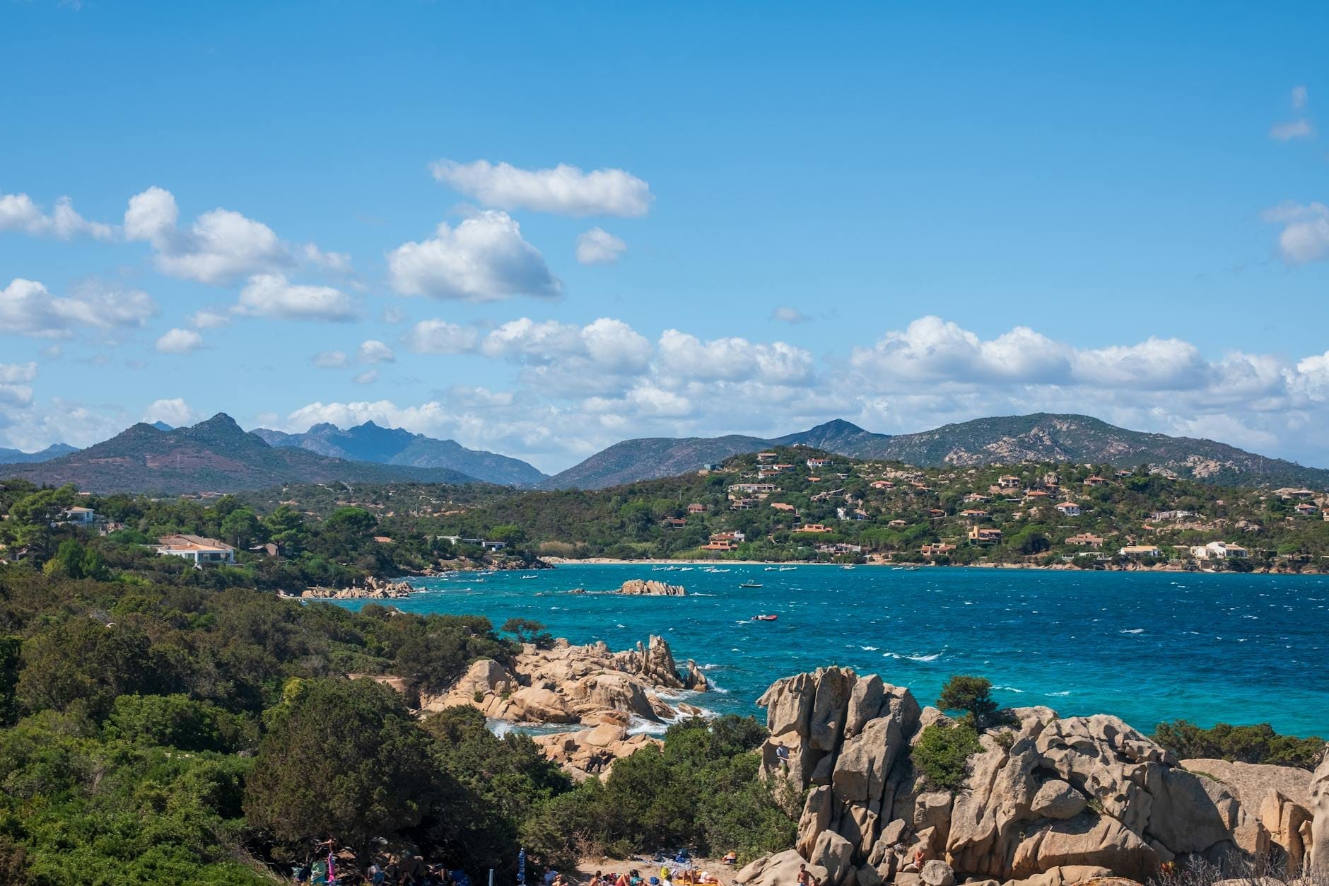 Küstenlandschaft auf Sardinien mit türkisfarbenem Meer, felsiger Küstenlinie, grünen Hügeln und fernen Bergen unter einem blauen Himmel mit vereinzelten Wolken.