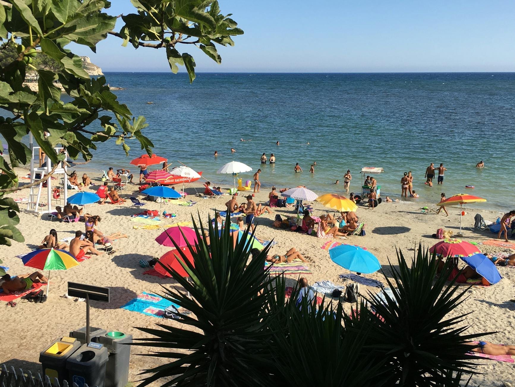 Belebter Sandstrand mit Familien, bunten Sonnenschirmen und spielenden Kindern am klaren blauen Wasser an einem sonnigen Tag.