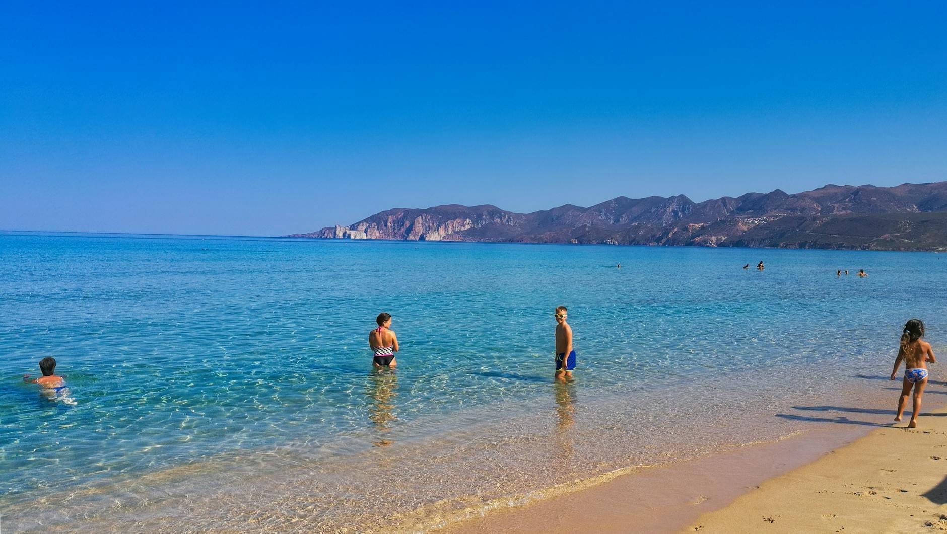 Kinder und Familien plantschen im flachen türkisfarbenen Wasser an einem Sandstrand mit Bergen im Hintergrund an einem sonnigen Tag auf Sardinien.
