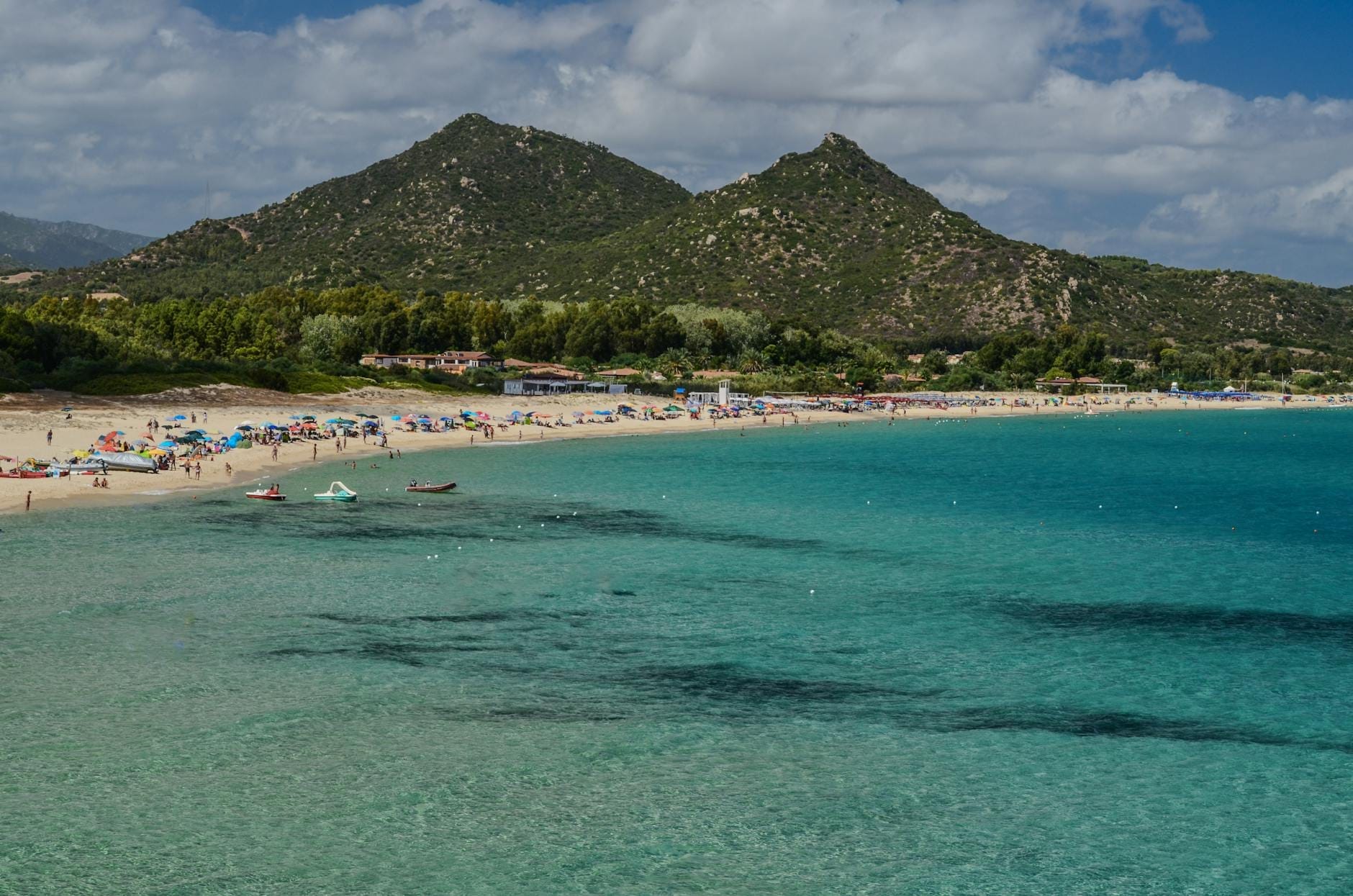 Ein breiter sardischer Strand mit türkisfarbenem Wasser, sanften Wellen, Sonnenschirmen, Familien und fernen grünen Hügeln unter einem teils bewölkten Himmel.