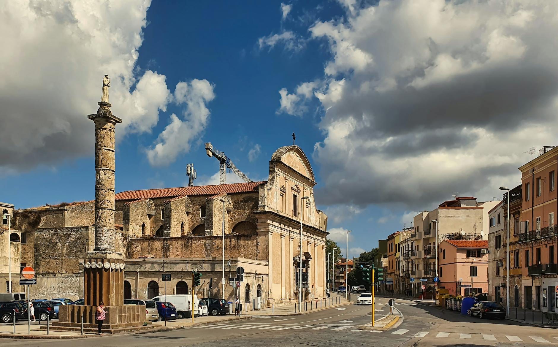 Ein weitläufiger Stadtplatz in Sassari, Sardinien, gesäumt von historischen Gebäuden und einer Kirche, mit einer markanten Säule und dramatischen Wolken darüber.