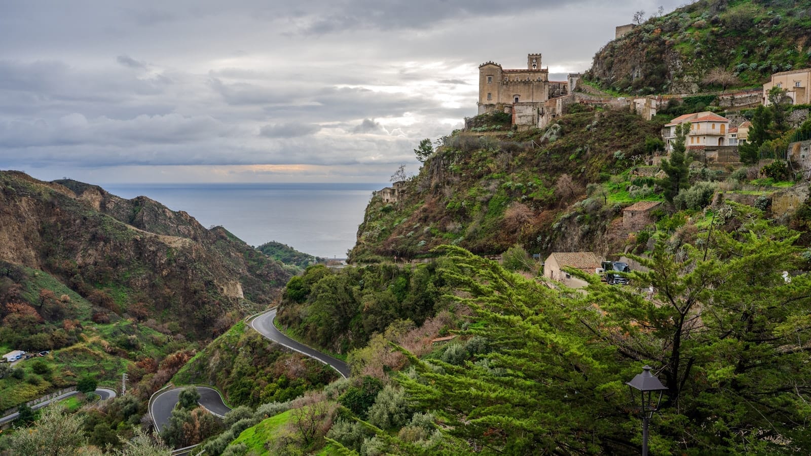 Das Bergdorf Savoca mit historischen Steingebäuden und üppig grünen Hügeln mit Blick auf das Ionische Meer an einem bewölkten Tag auf Sizilien.