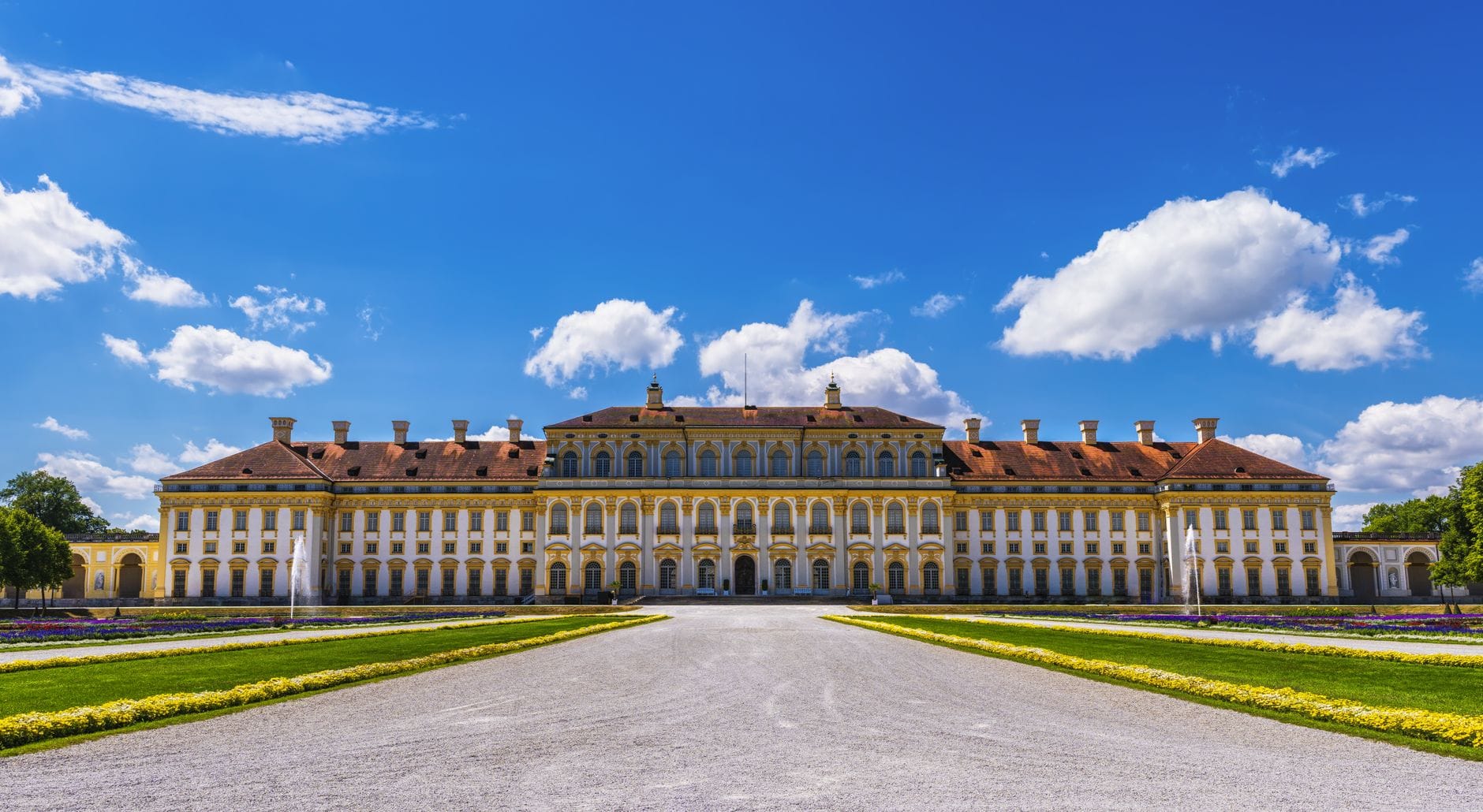 วิวมุมกว้างของพระราชวังบาโรกหลักของ Schloss Schleissheim ใต้ท้องฟ้าสีคราม แวดล้อมด้วยสวนจัดแต่งสวยงามและทางเดินโรยกรวดสุดหรูด้านหน้า