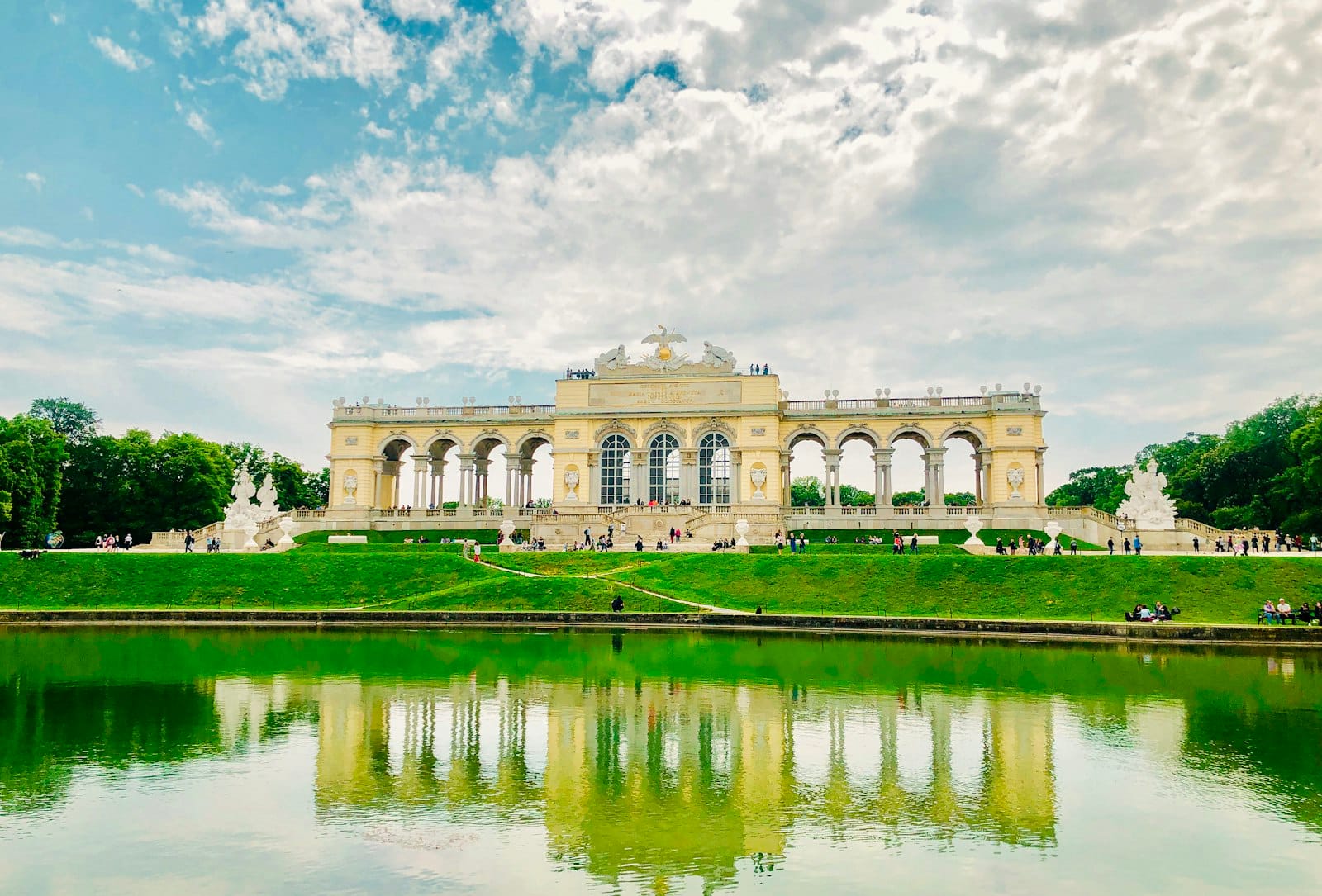 Weiter Blick auf die Kolonnade der Schönbrunn Gloriette, die sich in einem Teich spiegelt, mit dramatischen Wolken und sattem Grün an einem hellen Tag.