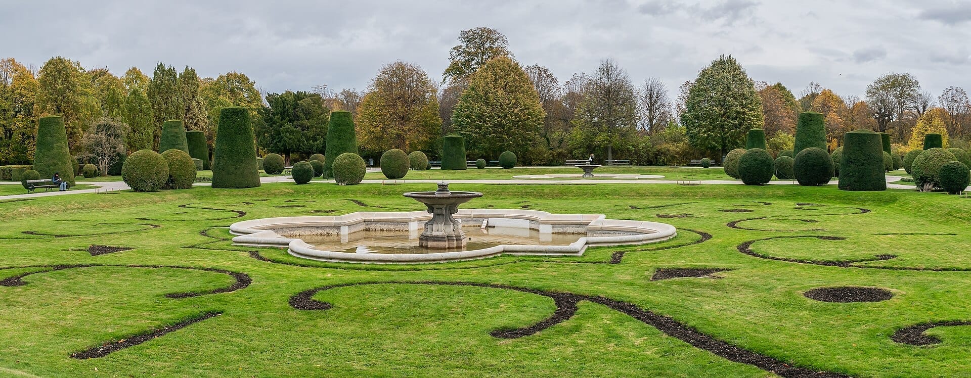 Symmetrisch geschnittene Hecken, kunstvoll gestaltete grüne Rasenflächen und ein zentraler Brunnen im Schlosspark Schönbrunn an einem bewölkten Herbsttag.