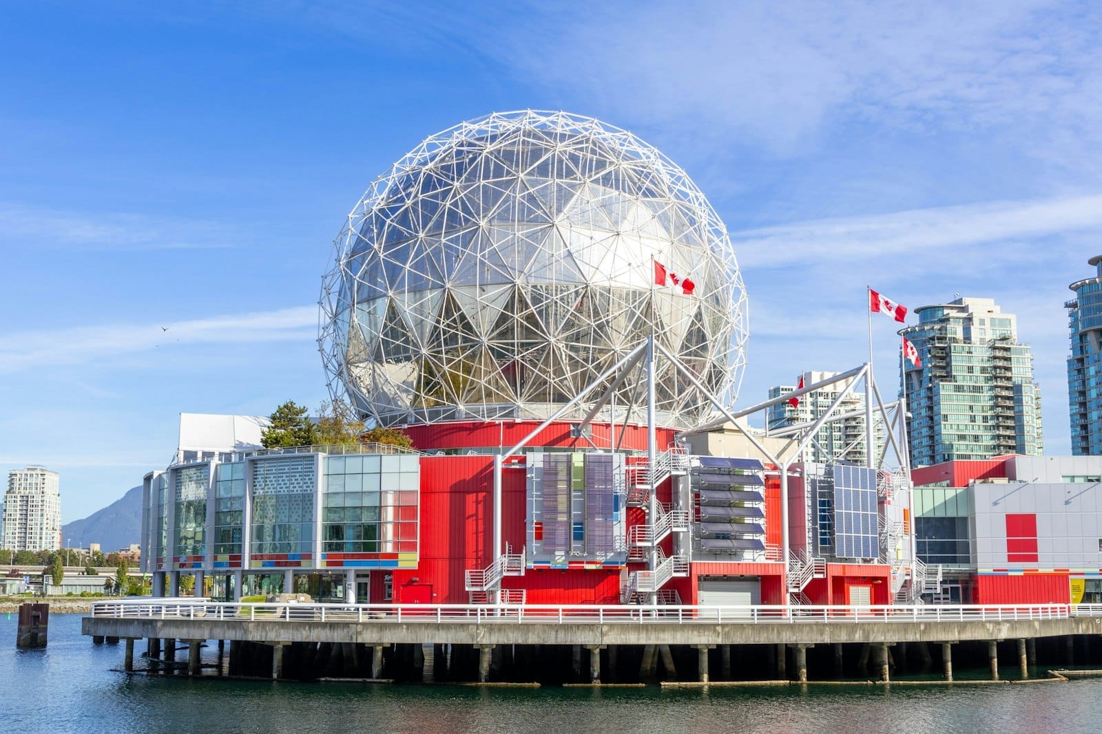 วิวภายนอกโดมจีโอเดสิกอันโดดเด่นของ Science World Vancouver พร้อมอาคารสีแดงและกระจก ธงชาติแคนาดา และริมน้ำ False Creek ใต้ท้องฟ้าสีฟ้า