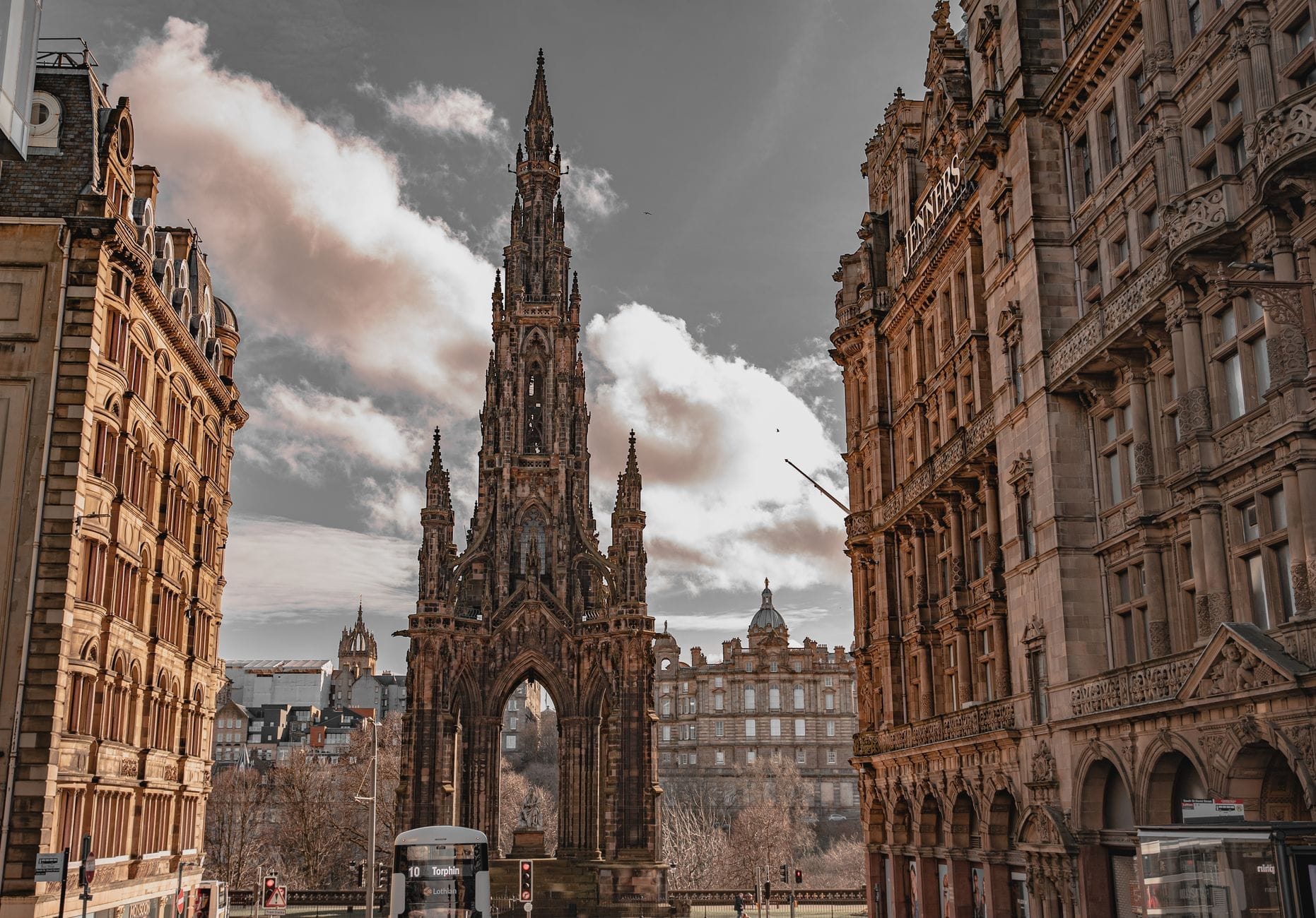 Das Scott Monument überragt die Princes Street Gardens in Edinburgh, eingerahmt von verzierten viktorianischen Gebäuden unter einem teilweise bewölkten Himmel mit Stadtverkehr darunter.