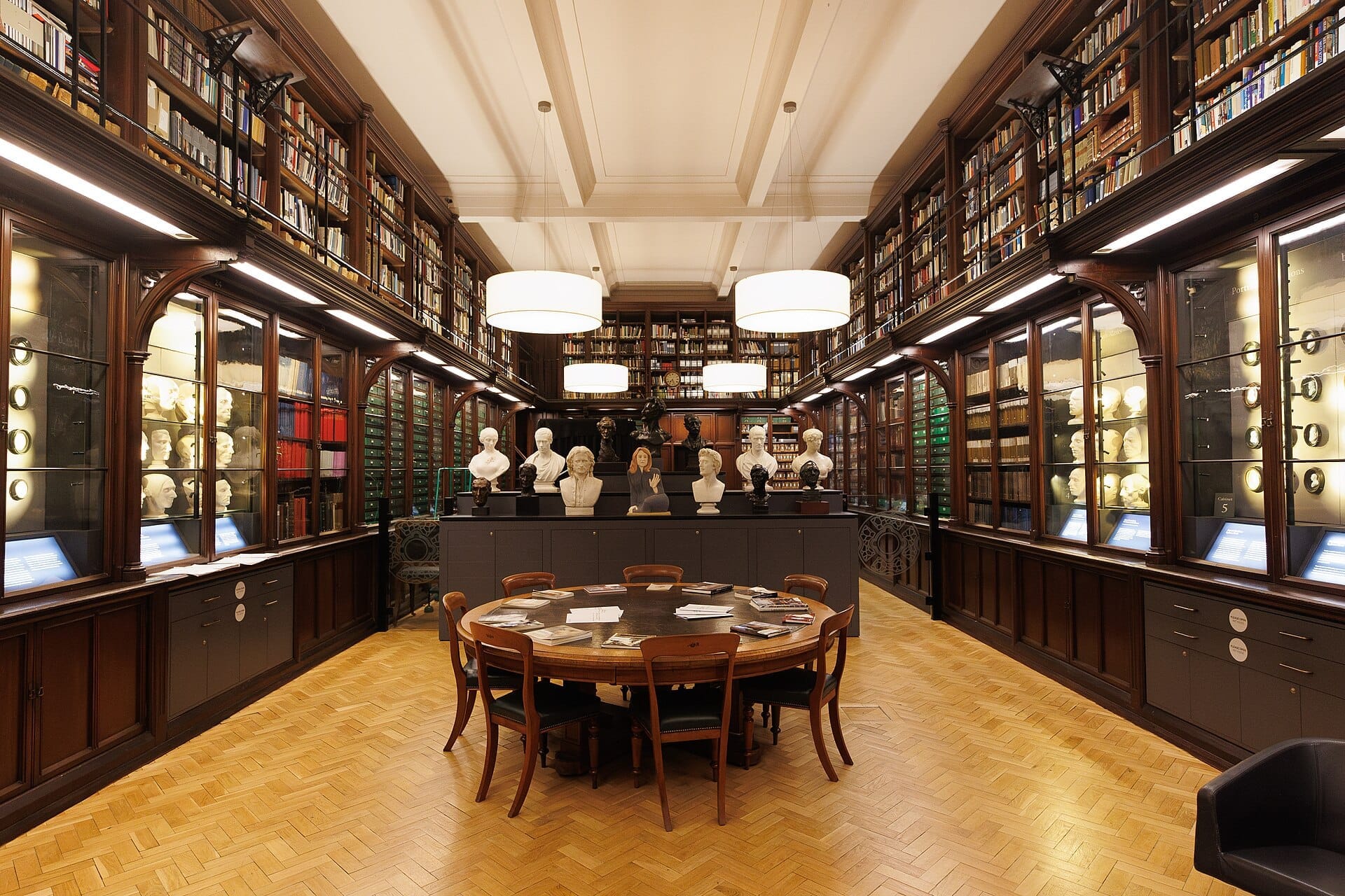 Der Lesesaal der Scottish National Portrait Gallery mit Holzbücherregalen, Marmorbusten, Glasvitrinen und einem runden Tisch unter warmem Licht.
