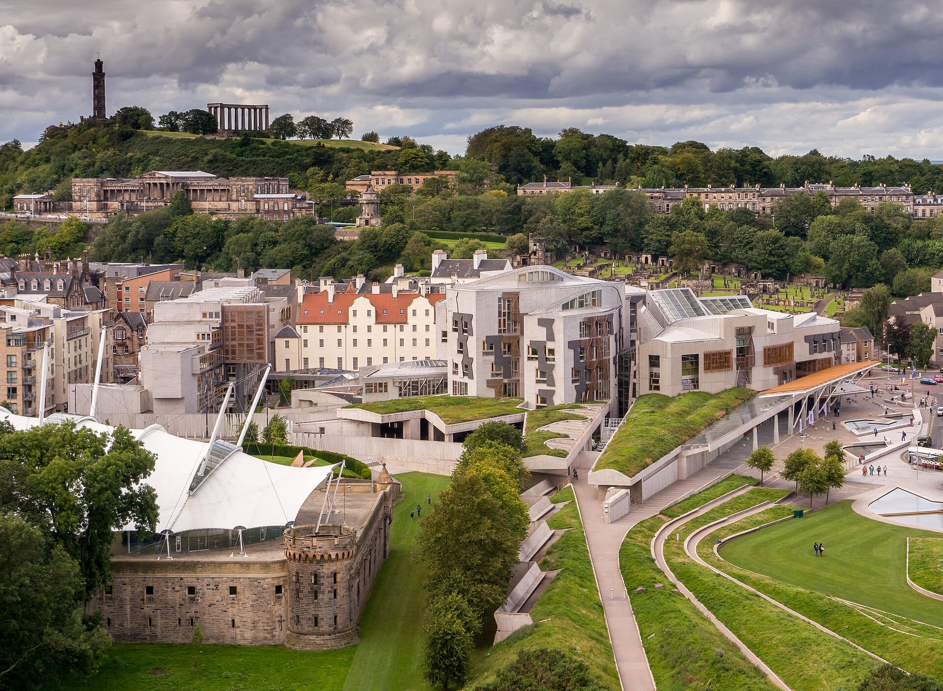 Eine weitläufige Luftaufnahme des modernen Schottischen Parlamentsgebäudes in Holyrood, Edinburgh, mit seiner markanten Architektur, angrenzenden historischen Gebäuden und grünen Hügeln.