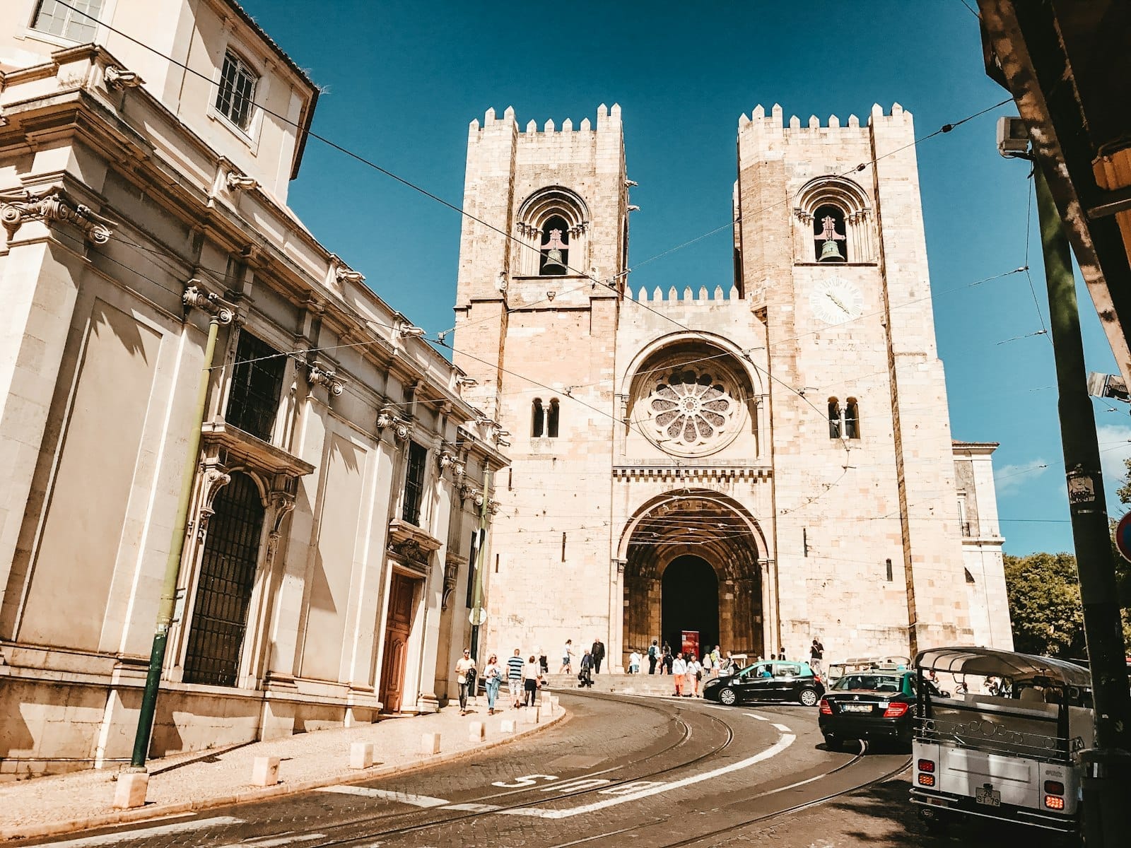 La Cattedrale Sé a Lisbona, con le sue due torri e il rosone, vista in una giornata di sole con persone e taxi davanti.
