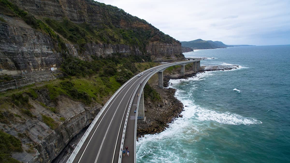 Vista aérea del Sea Cliff Bridge curvándose a lo largo del dramático litoral del Mar de Tasmania, con olas rompiendo abajo y acantilados verdes elevándose por encima.