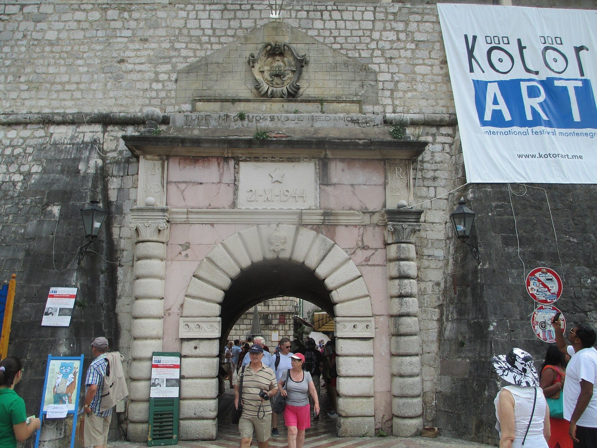 I visitatori attraversano lo storico arco in pietra della Porta del Mare di Kotor, adornato con rilievi e circondato dalle antiche mura della città vecchia.