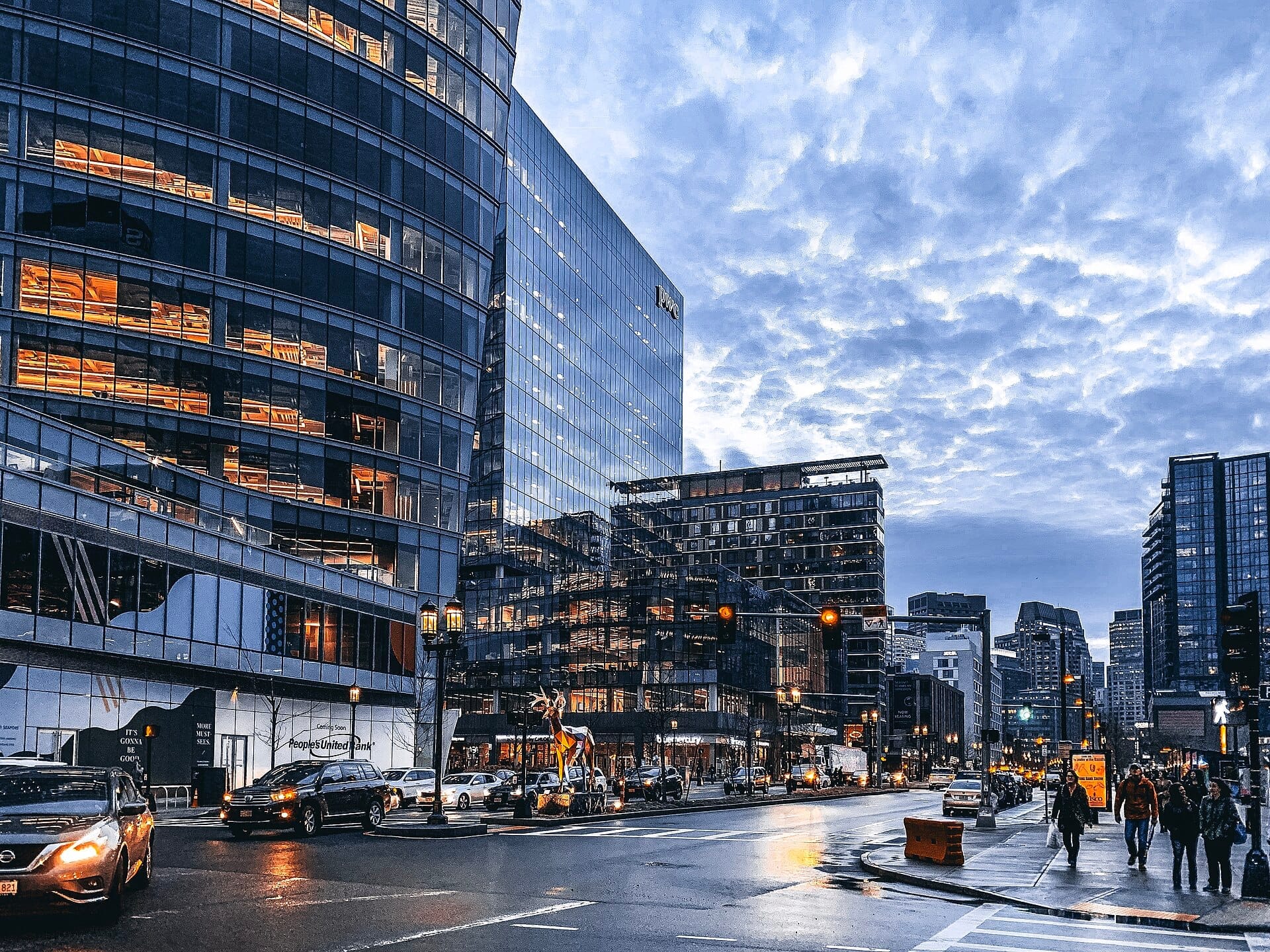 อาคารกระจกทันสมัยเรียงรายตามถนนที่คึกคักในย่าน Seaport District ของ Boston ยามพลบค่ำ พร้อมรถยนต์และคนเดินถนนที่สัญจรไปมาใต้ท้องฟ้าครึ้มที่สวยงาม