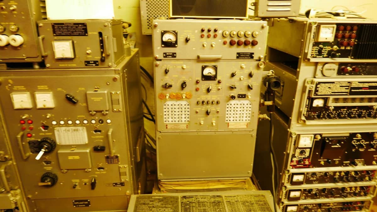 Reihen von Vintage-Kontrollpanelen und Ausrüstungen aus der Sowjetzeit im Inneren des Geheimen Sowjetischen Bunkers in Līgatne, mit Zifferblättern, Messgeräten und Schaltern unter gelbem Licht.