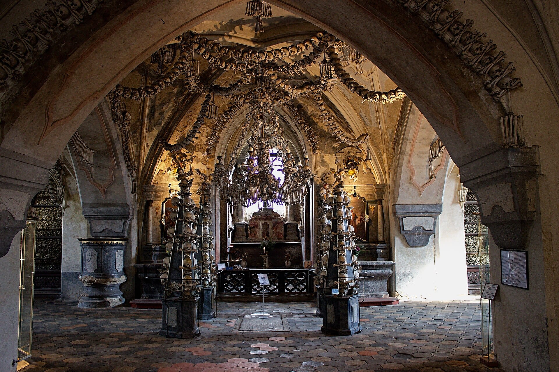 Interno dell'Ossario di Sedlec con archi gotici, lampadari e sculture adornati con ossa e teschi umani sotto una luce fioca.