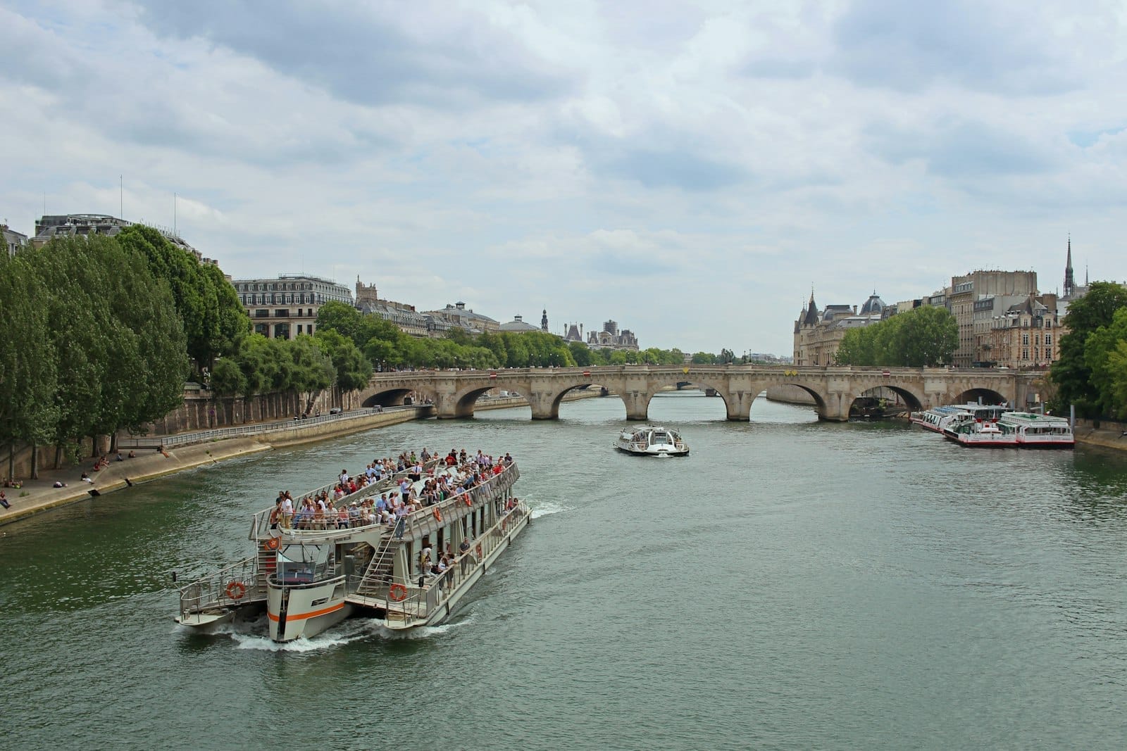 Un battello turistico pieno di passeggeri scivola lungo la Senna a Parigi, passando sotto storici ponti e lungo rive ombreggiate da alberi in una giornata luminosa.