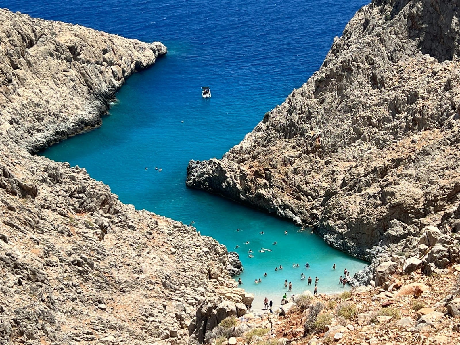 Vista aerea della baia di Seitan Limani con acque turchesi, ripide scogliere rocciose e persone che nuotano nella piccola spiaggia sabbiosa.
