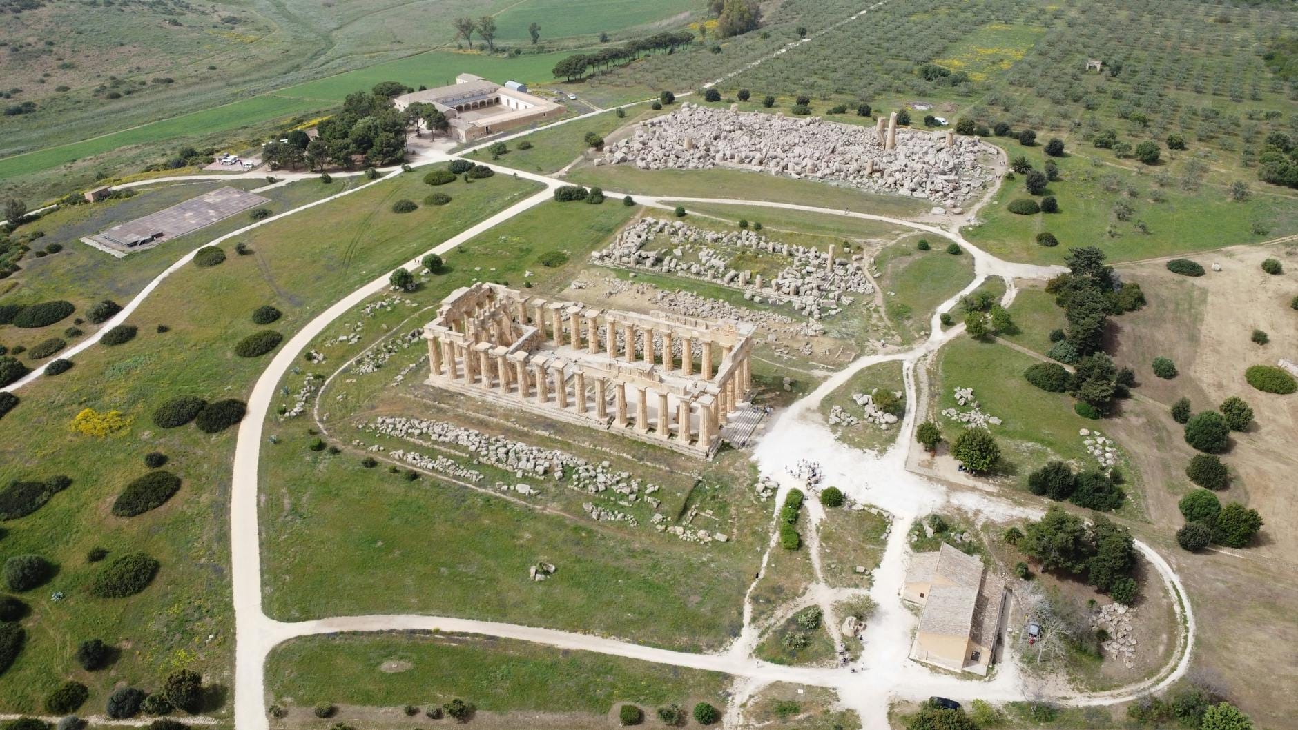 Luftaufnahme des Archäologischen Parks Selinunt mit gut erhaltenen griechischen Tempelruinen, Steinfundamenten, geschwungenen Wegen und grüner Landschaft unter klarem Himmel.