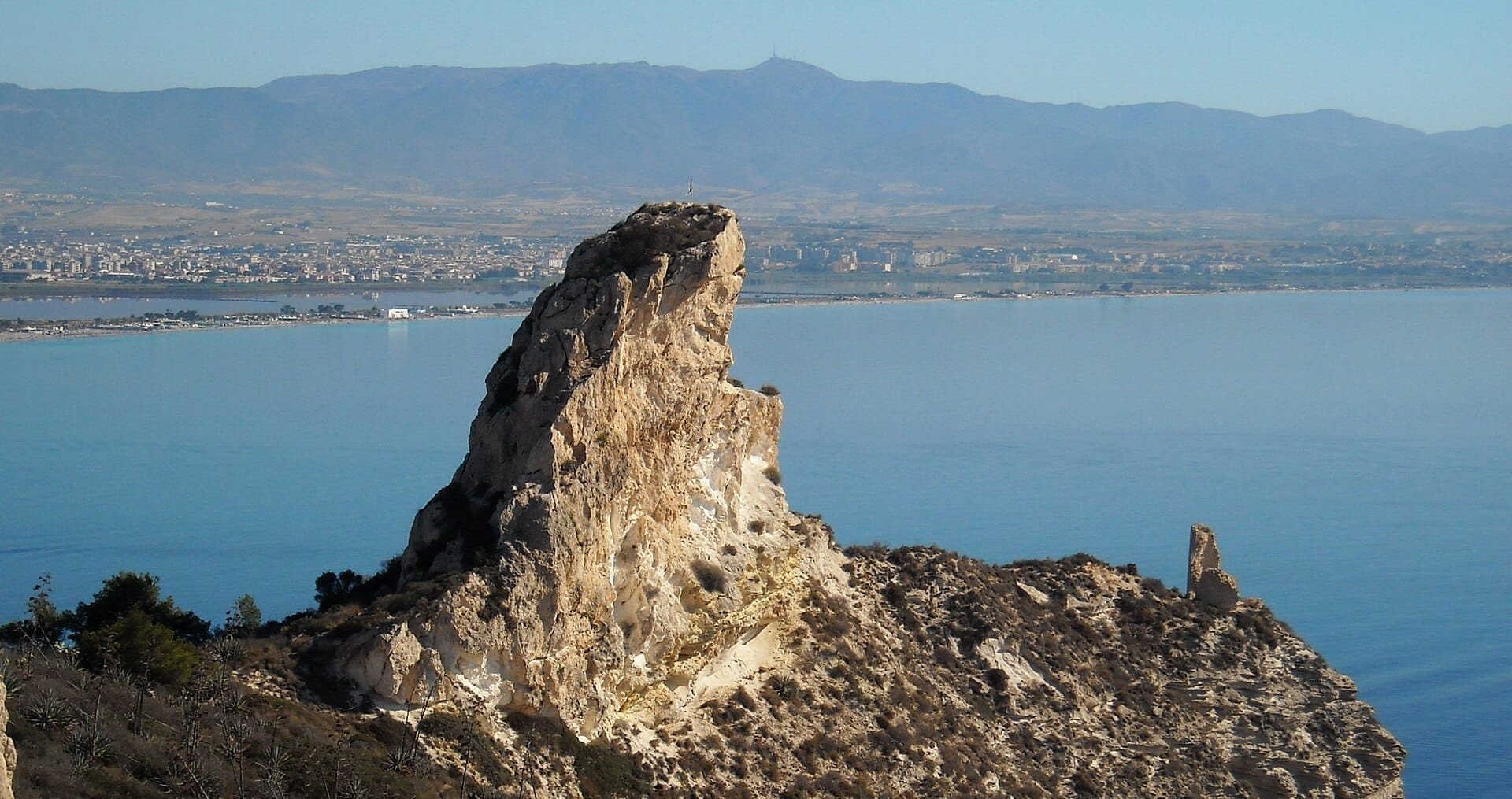 Markierter Kalksteinausläufer der Sella del Diavolo, der sich über die blaue Küste erhebt, mit der Stadt Cagliari und Bergen im Hintergrund.