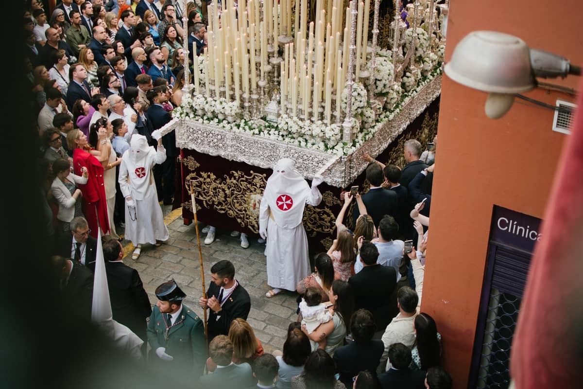 ขบวนแห่ Semana Santa ที่ประดับด้วยเทียนไขในถนนที่คับคั่งของเมือง Sevilla ล้อมรอบด้วยผู้คนในชุดคลุมและหมวกคลุมแบบดั้งเดิม