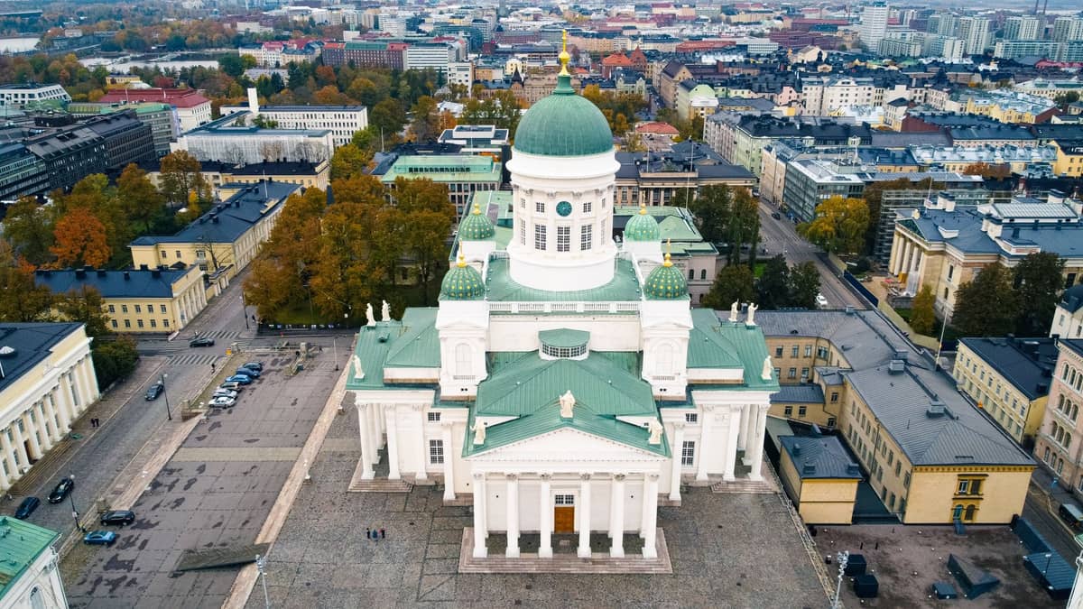 Vista aérea da Catedral de Helsinque dominando a Praça do Senado, cercada por edifícios neoclássicos governamentais e universitários, com o bairro de Kruununhaka ao fundo.