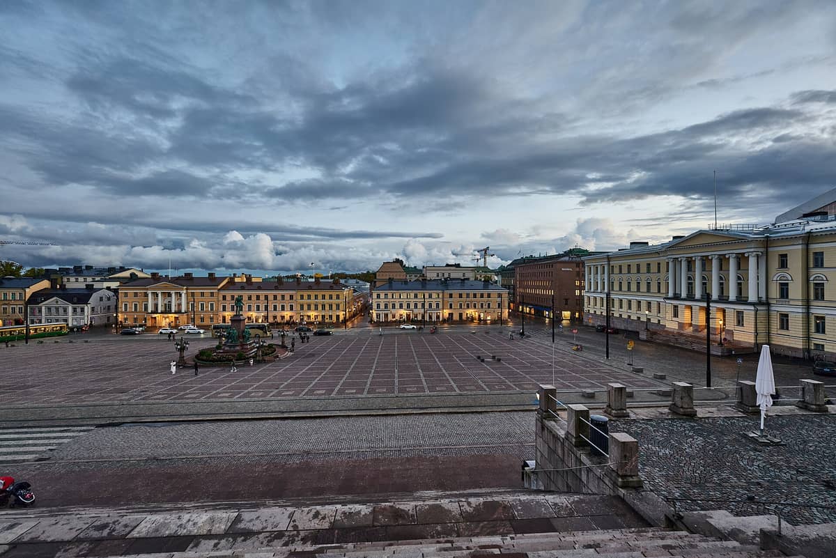 Vista ampla da Praça do Senado de Helsinque ao entardecer, com os edifícios neoclássicos, a praça central e a estátua central fotografados a partir das escadarias da catedral.
