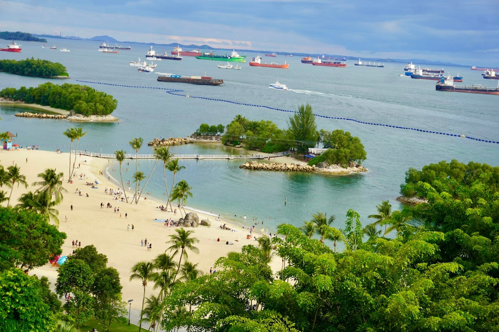 วิวพาโนรามาชายหาดเซ็นโตซากับต้นปาล์ม หาดทราย และเกาะเล็กนอกชายฝั่ง ผู้คนมากมายท่ามกลางเรือสินค้าไกลออกไปใต้ท้องฟ้าสีคราม
