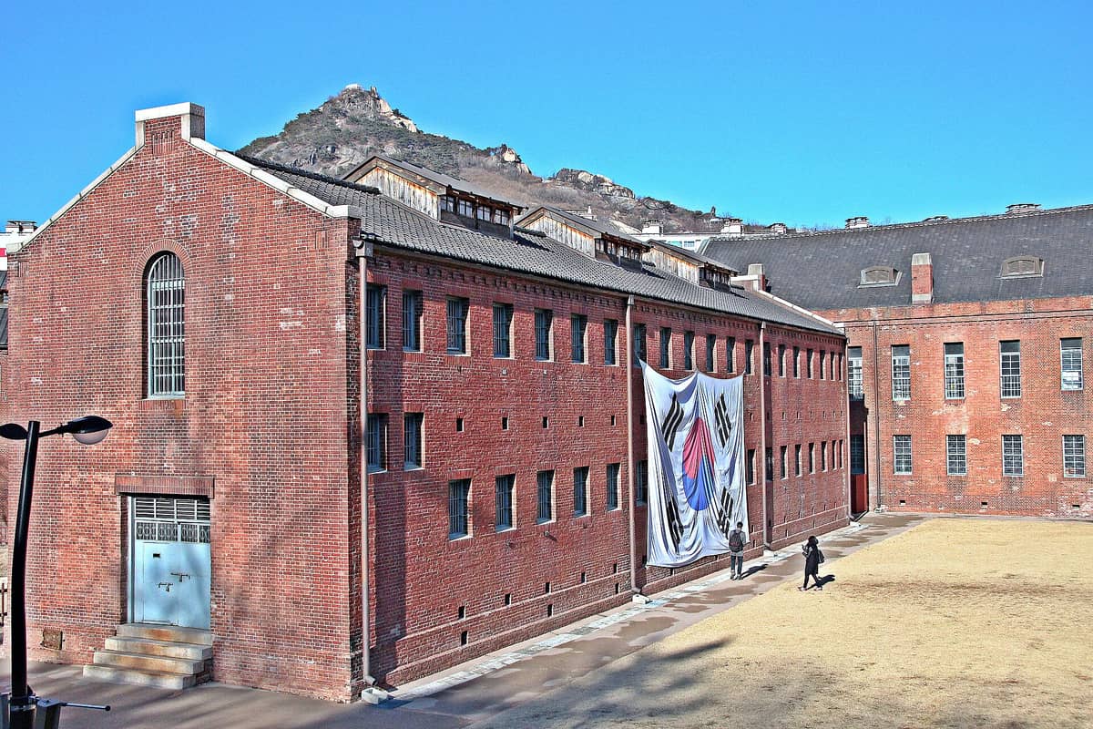Fachada do Museu Histórico da Prisão de Seodaemun com seu icônico prédio de tijolos vermelhos, janelas gradeadas e uma grande bandeira coreana, ao fundo um céu azul e montanhas.