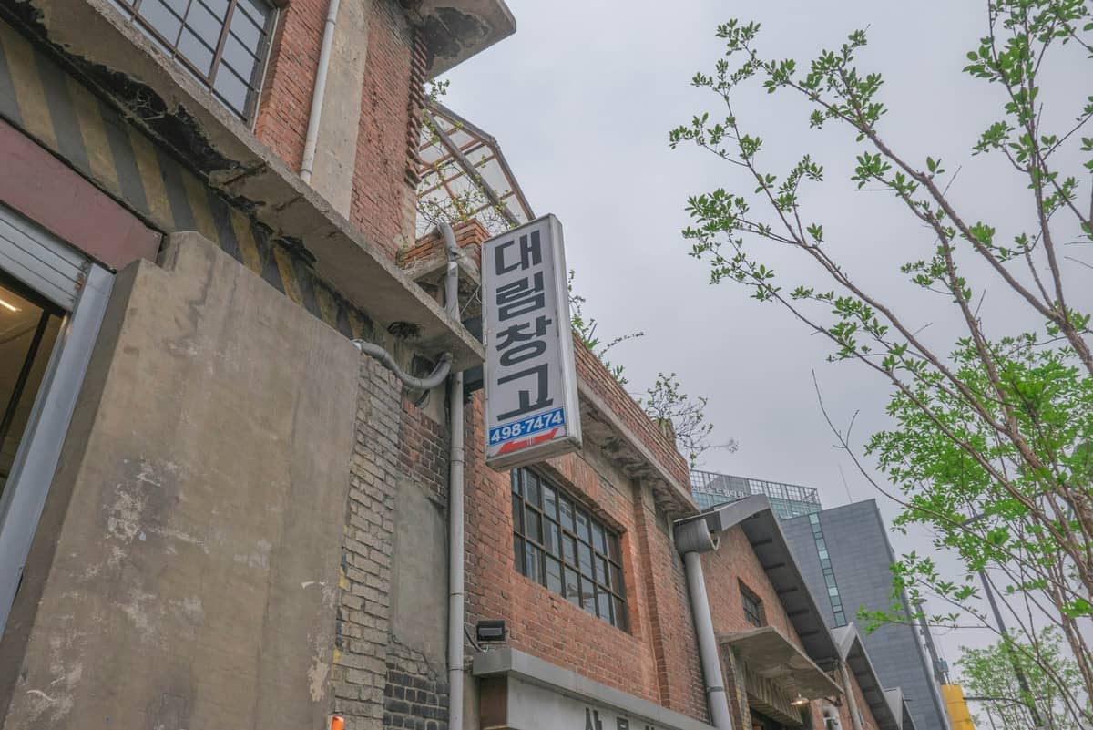 Edifício industrial de tijolo vermelho com uma placa em coreano e grandes janelas em Seongsu, Seul, com um céu nublado e galhos verdes frescos ao fundo.