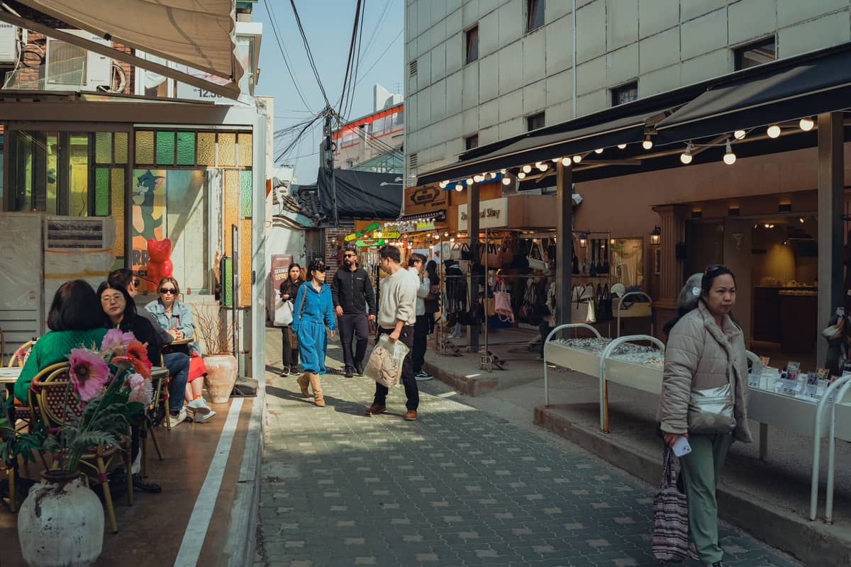 Movimentada cena de rua em Seul com pessoas sentadas do lado de fora de um café colorido e caminhando por lojas únicas em um vibrante beco.