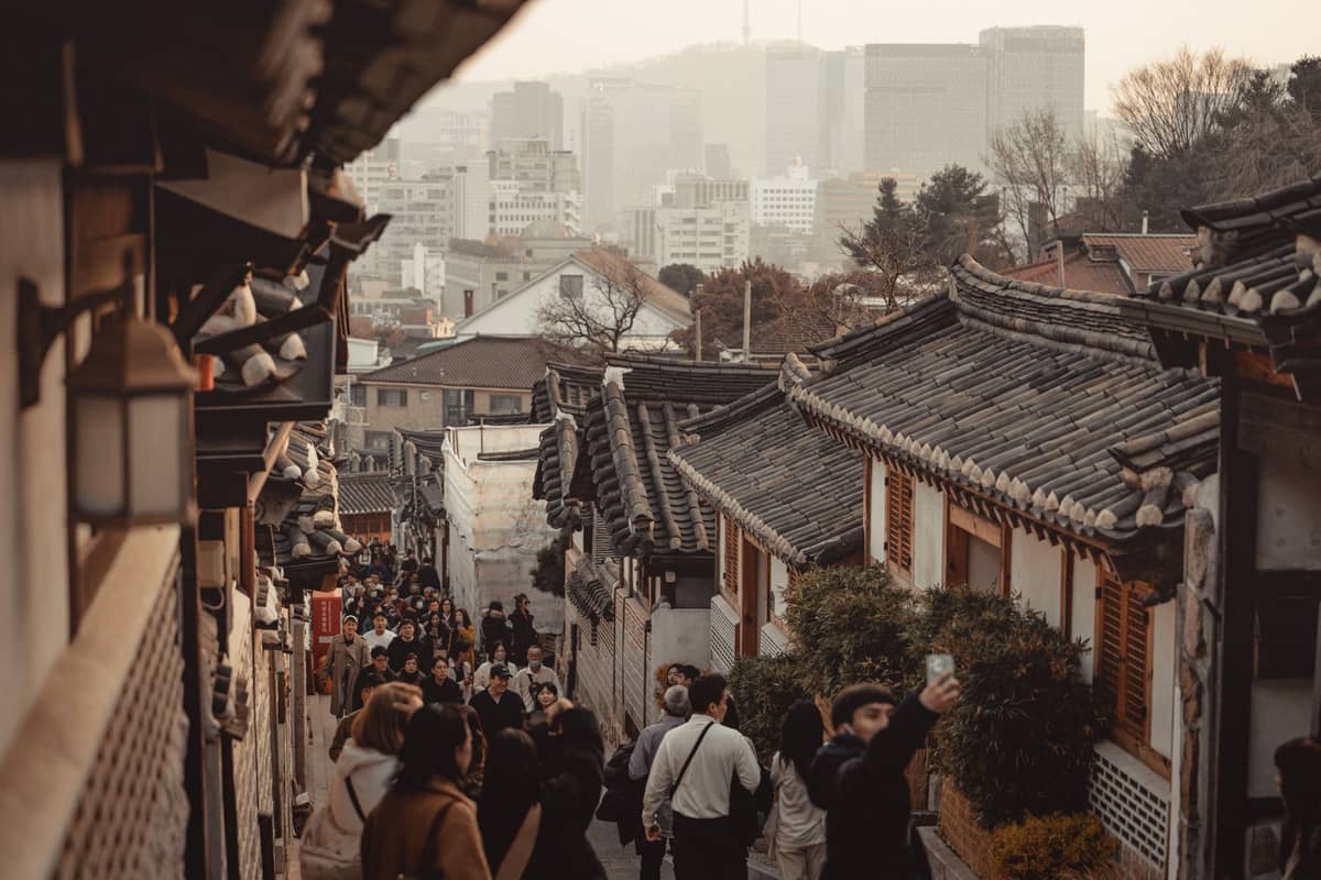 Rua movimentada em Bukchon Hanok Village com casas coreanas tradicionais, hanok, alinhadas em ambos os lados e edifícios modernos da cidade visíveis ao fundo enevoado.