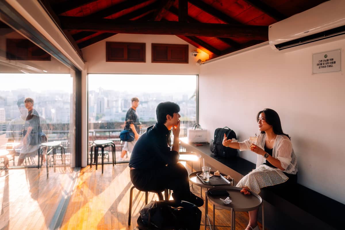 Interior de um café moderno em Seul com grandes janelas, vista para a cidade e pessoas curtindo café e sobremesas em um ambiente descontraído.