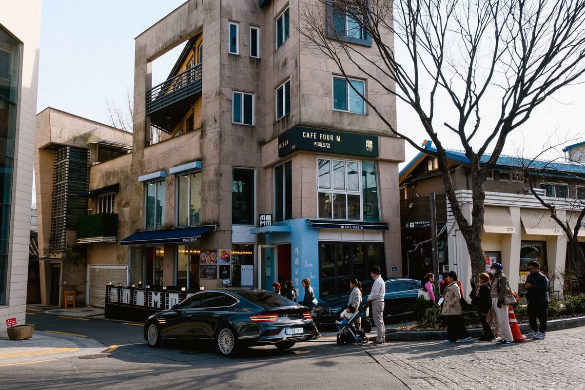 Moderno edifício de café com vários andares em uma esquina no bairro descolado de Seul, com pessoas esperando do lado de fora e carros estacionados ao longo da rua