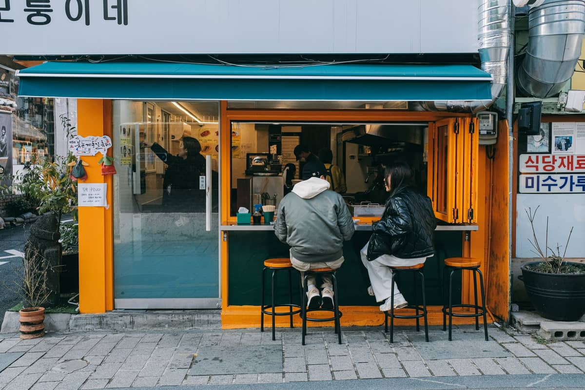 Duas pessoas sentadas no balcão aberto de um café em uma rua para pedestres em Seul, com detalhes coloridos e modernos e equipamentos de café visíveis no interior.