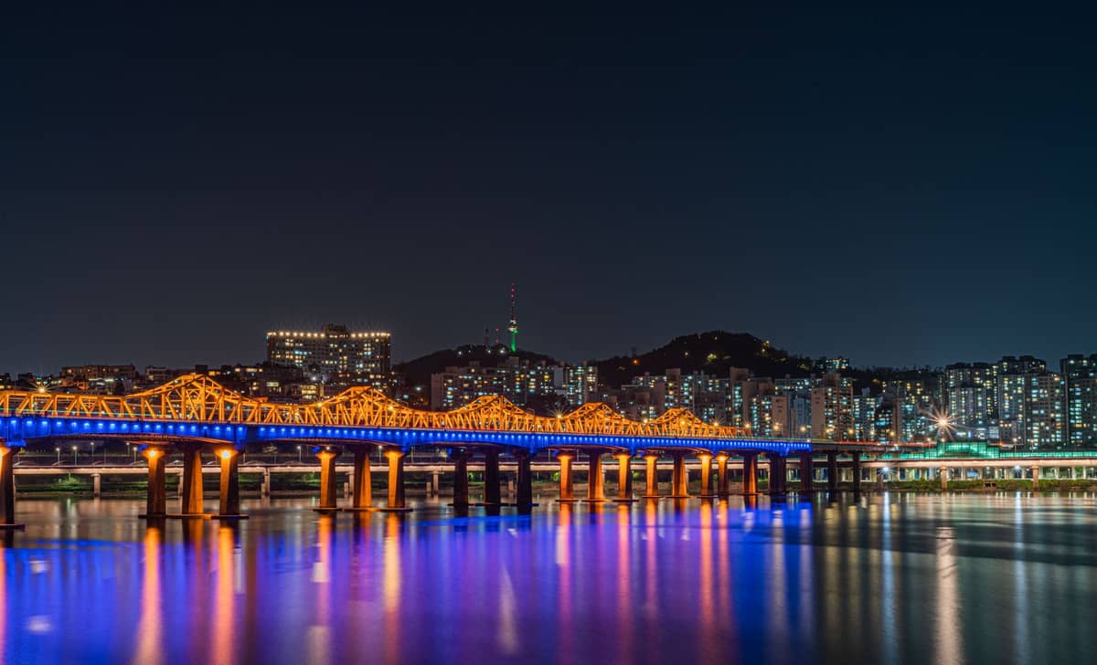 Ponte iluminada com cores sobre o rio Han à noite com o horizonte de Seul e a Torre Namsan Seoul ao fundo.