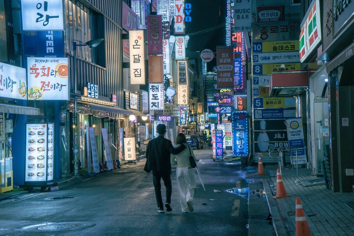 Um casal caminha de mãos dadas por uma rua iluminada com néons em Seul à noite, cercado por letreiros coloridos e uma atmosfera urbana romântica.