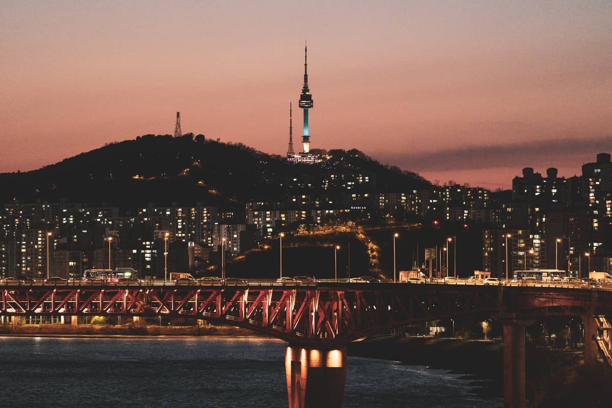 A Torre Namsan de Seul ergue-se iluminada no topo de uma colina ao entardecer, com o horizonte da cidade e uma ponte sobre o Rio Han em primeiro plano.