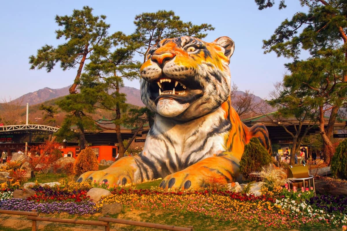 Grande estátua colorida de tigre na entrada do Seoul Grand Park Zoo, cercada por flores, árvores e montanhas na primavera.