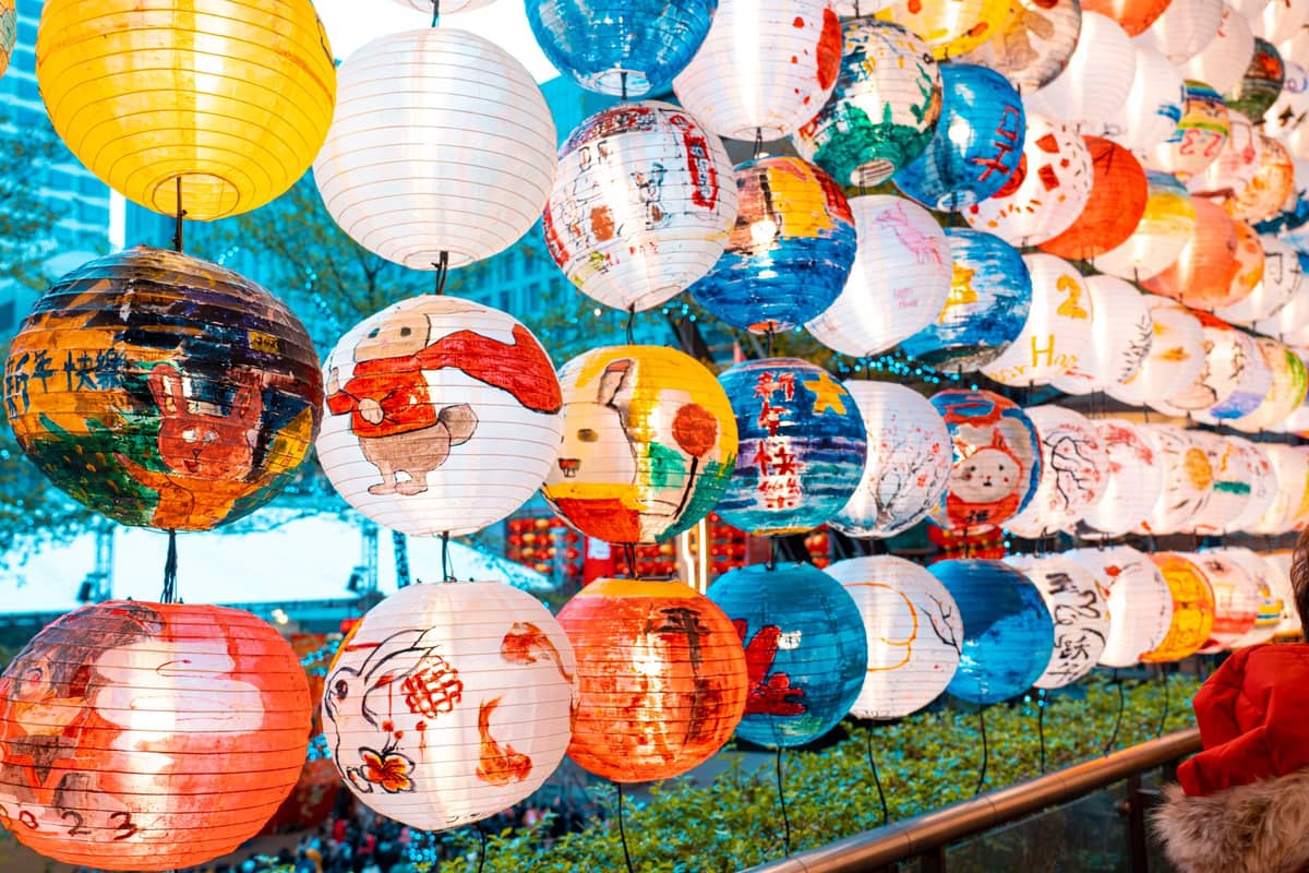 Lanternas de papel pintadas à mão em cores vibrantes penduradas em fileiras, provavelmente em um festival, com alguns edifícios da cidade e árvores visíveis ao fundo.